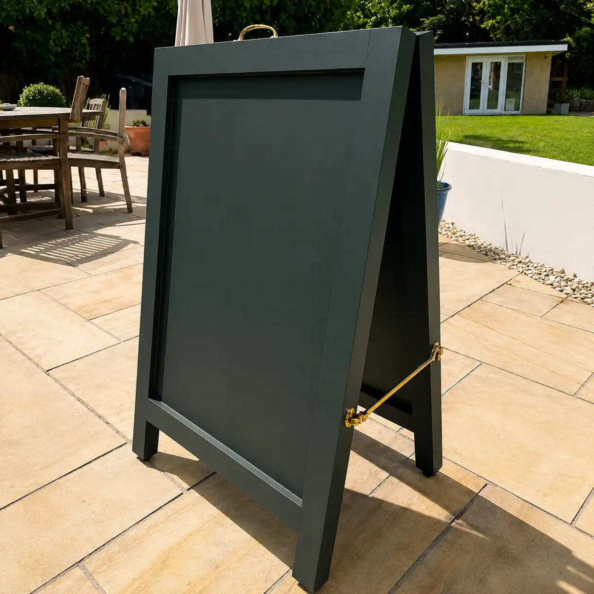 A dark greenA-frame sidewalk sign with a gold metal handle and brass hardware on a tiled outdoor patio.