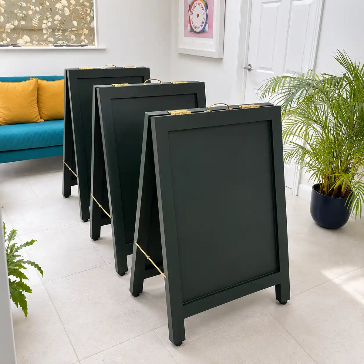 Three dark green A-frame pavement boards with brass  metal handles are lined up on a light-colored tiled floor in a bright, modern room with a teal sofa and potted plants.